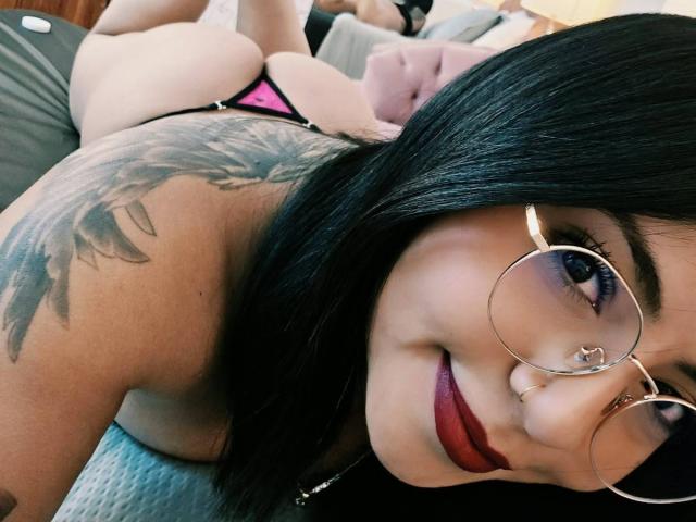 IsaLawrence, avec ses cheveux noirs, ses lunettes, son anneau au nez et son rouge à lèvres rouge, est allongée sur le ventre, montrant un tatouage sur son épaule, portant un string rose et noir, et souriant à la caméra..