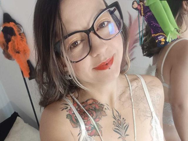 ArianaBlonde with glasses, red lipstick, and a nose ring, smiling at the camera. She has tattoos on her chest and is wearing a white lace top.