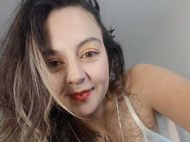 ArianaBlonde with light brown, wavy hair, red lipstick, and tattooed chest, wearing a white lace top, smiling at the camera. Her eyes are golden and she's looking up.