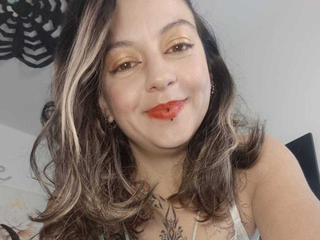 ArianaBlonde with wavy brown hair, orange eyeshadow, red lipstick with a nose ring, and a tattoo on her chest, smiling at the camera. Boobs visible, arms slightly out.