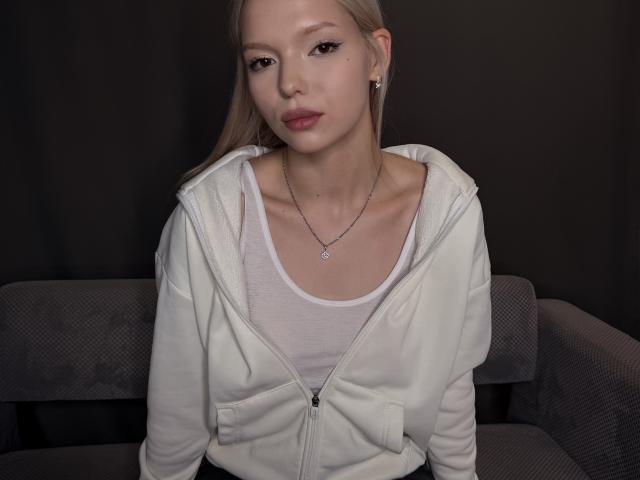 Willodean Almonte mit blonden Haaren, einem weißen Kapuzenpullover und einem weißen Tanktop, der kleine Brüste zeigt, trägt eine Halskette und sitzt auf einem grauen Sofa in einem dunklen Raum..