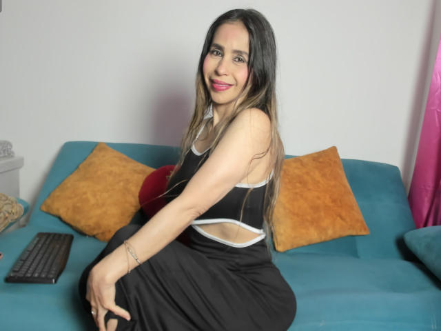 VictoriaJhonnes with long black hair, smiling, sitting on a blue sofa, wearing a black and white top, black skirt, showing her ass, with orange pillows and a remote.
