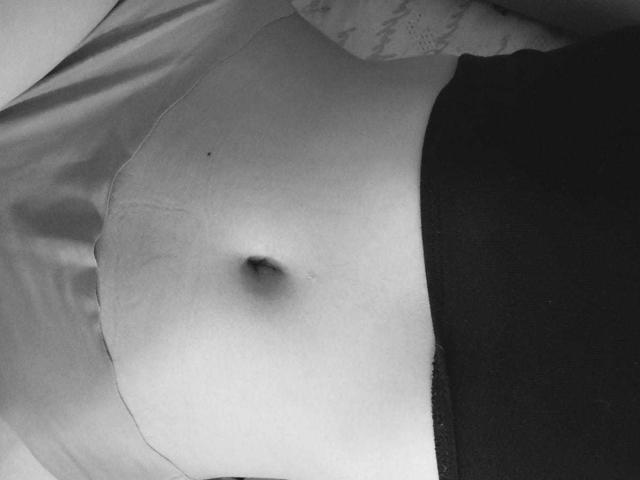 CherryBuchel's bare, light-skinned belly with a small navel, wearing black pants, lying on a bed, in a black-and-white photo.