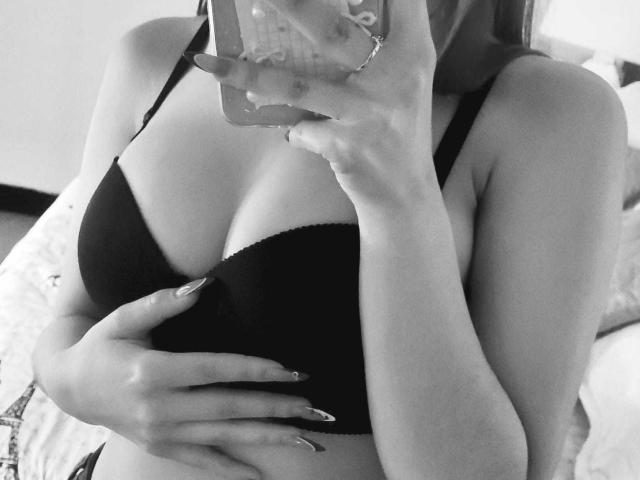 CherryBuchel with large, dark, underboob-exposing tits, wearing a black bra, holding a phone in front of her face, fingers with long, painted nails, in a black-and-white selfie.