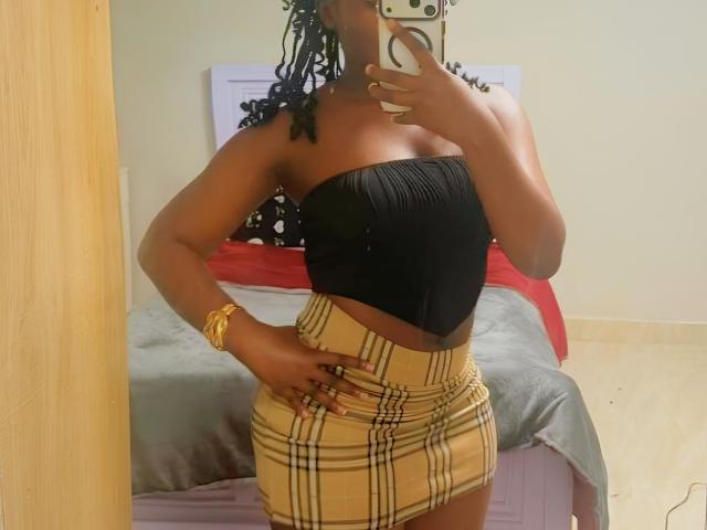 PriyTitty with dark skin, black strapless top, plaid high-waist skirt, gold bracelet, and blue-dyed braids, taking a mirror selfie with hand on hip, showing her ass.