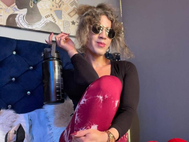 RossSofiia with curly gray hair, wearing round sunglasses and black top, holding a red heart-shaped pillow, right arm raised, black water bottle on blue tufted headboard.