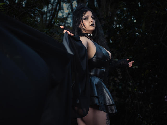 LilithAmoth, vêtue d'une robe en cuir noir qui révèle ses grandes seins, tenant un manteau noir, avec un maquillage sombre, de style gothique, sur un fond de forêt, avec de longs cheveux noirs, un collier ras du cou, debout avec assurance..