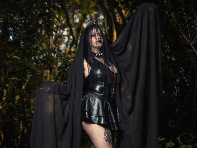 LilithAmoth, vêtue d'une robe en cuir noir, avec un voile noir, une couronne et des bas noirs, tenant le voile ouvert, montrant ses seins et sa cuisse tatouée, debout dans une forêt, portant du rouge à lèvres foncé..