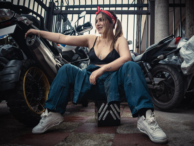 AriaRoa, with tits in a black top, sitting on a motorcycle box, wearing a red bandana, blue jeans, and white sneakers, surrounded by motorcycles in a garage.