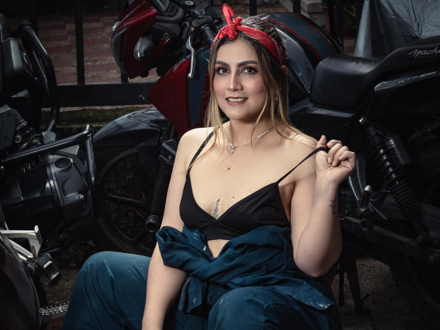 AriaRoa with small tits, wearing a black bra, denim overalls, and red headband, sitting by black motorcycles, holding bra strap, smiling, with a tattoo under her right breast.