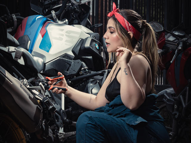 AriaRoa with tits in a black tank top, red bandana, and denim overalls, tinkering with a motorcycle, her fingers on the handlebars, looking focused. Motorcycle in front.