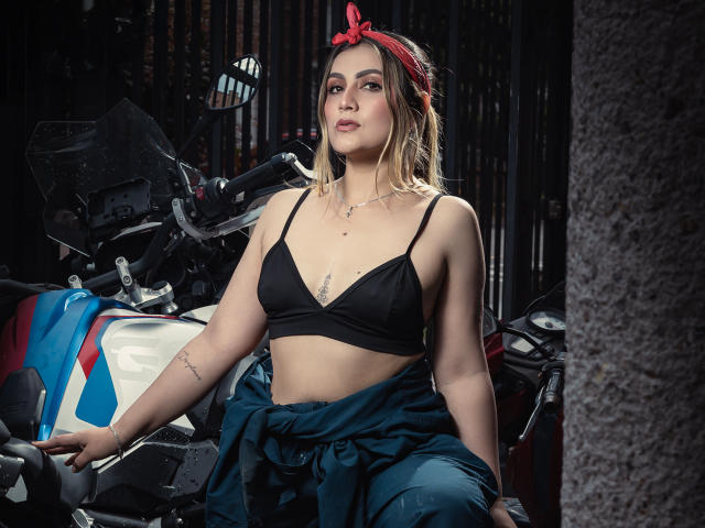 AriaRoa with small tits, wearing a black bra and red bandana, sits on a motorcycle, blue pants tied at waist, tattoo on right arm, looking confident.