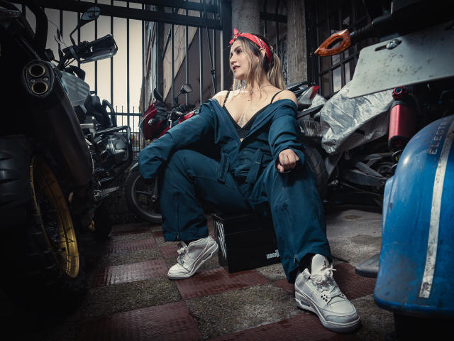 AriaRoa sits in a dirty garage, wearing an off-shoulder blue jumpsuit, white sneakers, and a red bandana, exposing her tits, with motorcycles and a blue tank around her.