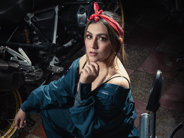 AriaRoa with a red bandana, blue overalls, and exposed shoulder, sitting in a dark, dirty garage, looking up with a thoughtful expression, hand near her chin.