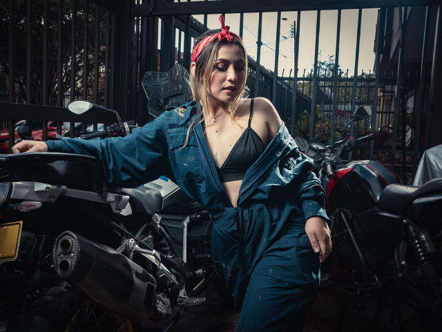 AriaRoa with blonde hair, red bandana, and blue overalls, exposing black bra and tits, leaning on black motorcycle in dark, gritty garage, looking down with a serious expression.