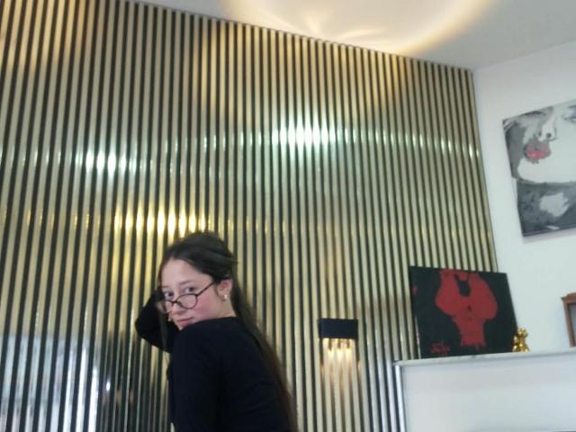 AmberBelle with glasses, long brown hair, black top, looking over shoulder, standing in front of shiny vertical gold wall, slight smile, white wall with art in background.