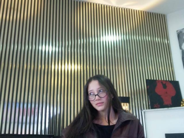 AmberBelle with long brown hair, glasses, and a brown coat, standing in front of a shiny, gold-striped wall, looking slightly to the side with a subtle smile.