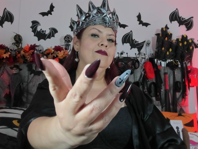 VanesaMoka with dark purple nails, wearing a silver spiked crown, black leather, and purple lipstick, showing her hand with a black and white nail design, in a Halloween-themed room.