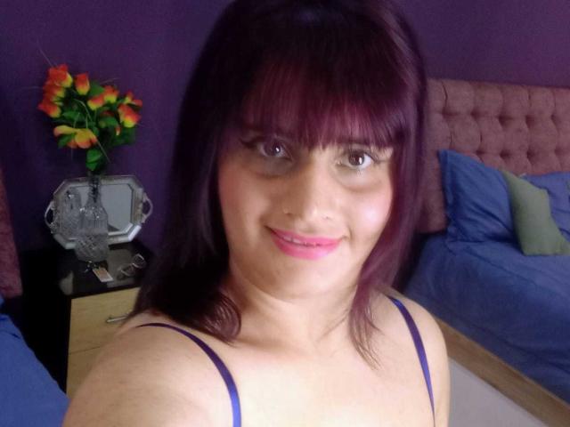 VivianFranco with purple hair and bangs, smiling, pink lipstick, wearing a purple spaghetti strap top, showing her tits, in a bedroom with blue beds and a vase of roses.