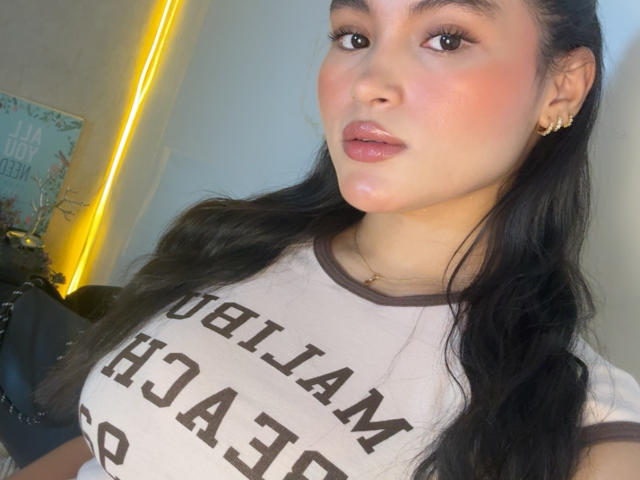 MiiaPalmer a de grandes seins brillants, vêtue d'une chemise moulante, avec de longs cheveux noirs, des lèvres roses brillantes et un boucle d'oreille dorée, regardant l'appareil photo avec une expression légèrement boudeuse..
