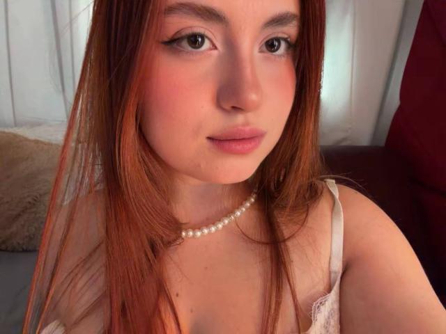VicluX a de longs cheveux roux, un collier de perles et un haut en dentelle blanche, avec un maquillage discret et des lèvres roses. Elle regarde l'appareil photo avec un léger sourire..