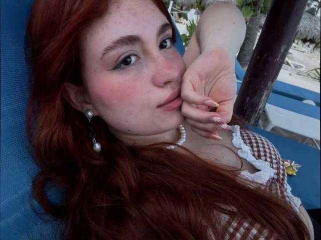 VicluX, com cabelo ruivo, sardas e brincos de pérola, com o queixo apoiado na mão, mostrando unhas longas, vestindo uma blusa xadrez, olhando para a câmera com um leve sorriso..