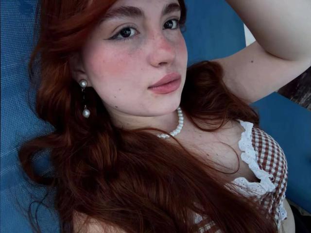 VicluX, com cabelo ruivo, vestindo um sutiã xadrez com renda branca, um colar de pérolas e brincos, tem sardas, lábios carnudos e um leve rubor, olhando para cima com um braço relaxado..