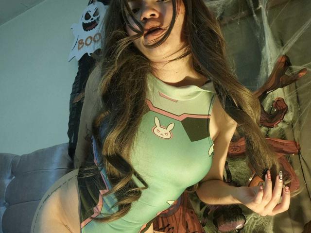 YourLuiseFantasy with long brown hair, green bodysuit, bunny patch, and dark lipstick, showing her tits, nails painted black, standing in a spooky room with fake spider webs.