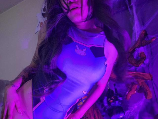 YourLuiseFantasy with long, wavy hair, wearing a tight, purple, bunny-patterned bodysuit, purple light, visible tattoos, holding a book, and a cute black cat toy in the background.
