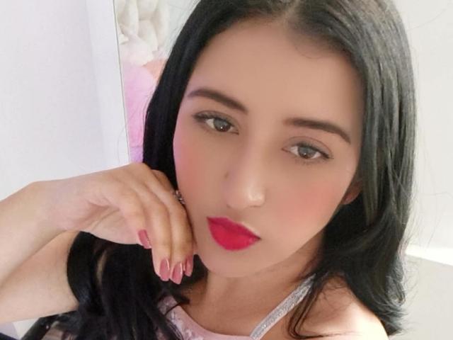 NicollJeans has long black hair, pink lipstick, and is looking at the camera with a slight smile. Her hand is near her face with pink nails.