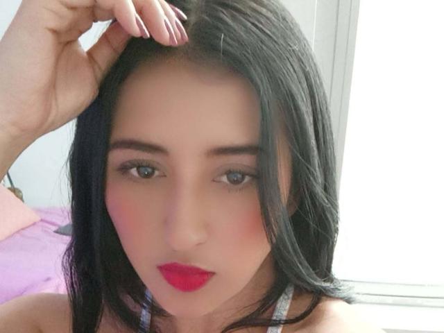 NicollJeans has pale skin, black hair, red lipstick, and is looking at the camera with a slight blush. Her hand is touching her head. She's wearing a white top.