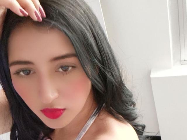 NicollJeans has black hair, red lipstick, and a silver necklace. She's touching her head with manicured pink nails. Her blue eyes are slightly downcast.