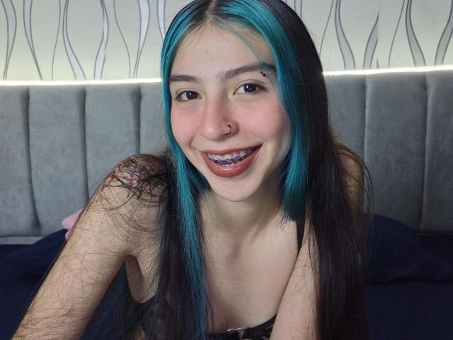 AbbyHillsI with blue-black hair, nose ring, and tooth jewelry, smiling with visible metal teeth, topless, showing small breasts, tattoo on right shoulder, on a bed.