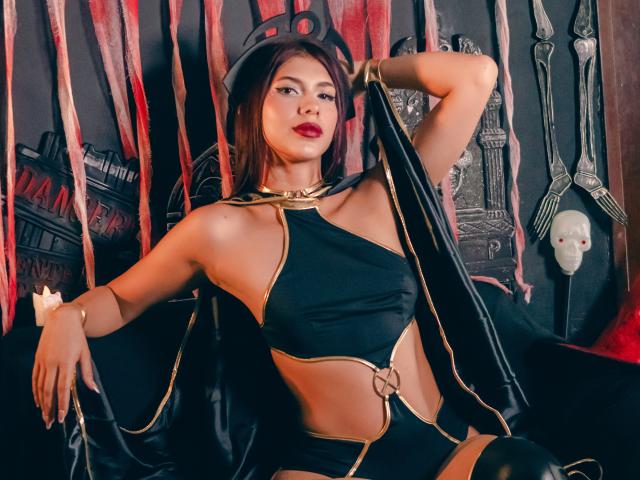 DanikaSerov with dark brown hair, wearing black and gold gothic lingerie, revealing her tits and pussy with a ring on her crotch, arm raised, red lipstick, confident pose.
