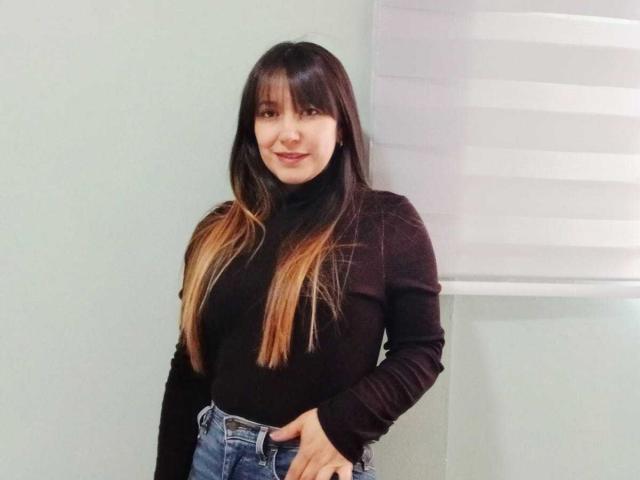 Roxanne Blair, avec de longs cheveux bruns dégradés en blond, un pull noir à col roulé et un jean bleu, debout, une main dans la poche, souriant doucement..