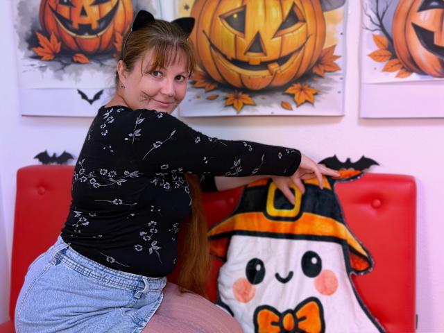 OVanessaHotly with long brown hair, black cat ears, black floral top, blue shorts, sitting on red couch, touching orange witch hat on cute white ghost pillow.