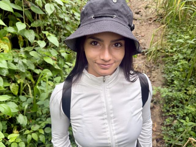 KimmyK with medium brown skin, black hair, wearing a grey bucket hat and white zip-up top, smiling, has small tits, standing in a green, leafy, outdoor area.