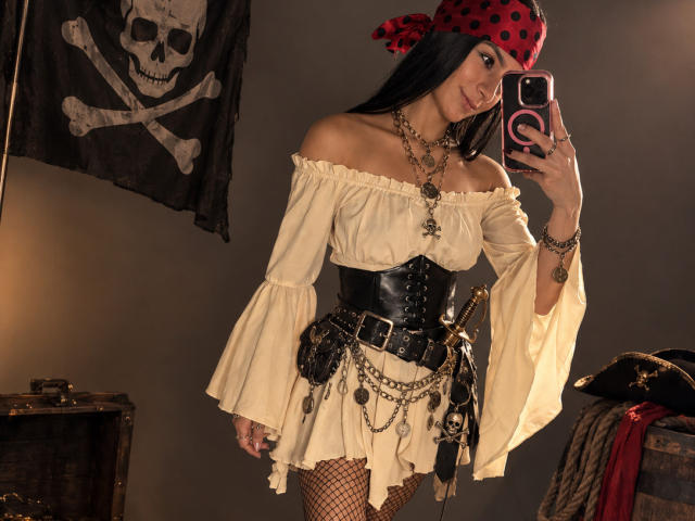 KimmyK with black hair, pirate hat, off-shoulder cream dress, black corset, fishnet stockings, multiple chains, and a skull flag, taking a mirror selfie, showing her tits.