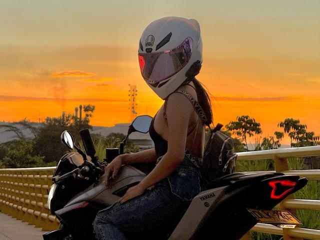 KimmyK with white helmet, black tank top, and blue jeans, sitting on a black Yamaha motorcycle, sunset in background, red light on helmet, holding motorcycle handle.