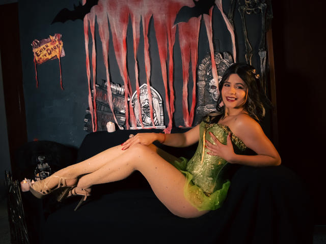 Sheyla Spooner con un corpiño verde de encaje, tul verde y tacones altos, sentada en un sofá negro, con las piernas cruzadas, sonriendo, cabello oscuro, piel pálida, pequeños senos, sin ropa interior, en un ambiente temático de Halloween..