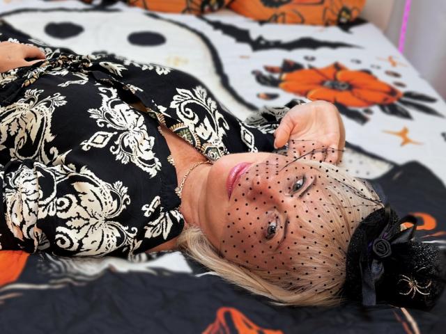 LaurenWishy lying on a bed, wearing a black and white floral dress, black lace hat with net, and showing off her blue eyes, with blonde hair and pink lipstick.
