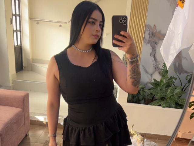 LilithAmoth, avec de longs cheveux noirs, portant une robe noire sans manches, un collier de perles et un bracelet, tenant un téléphone, montrant un tatouage sur son bras droit, dans un selfie pris devant un miroir..