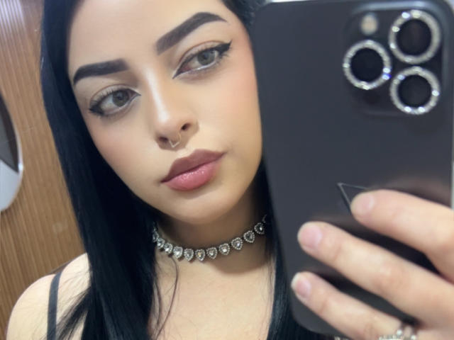 LilithAmoth a les cheveux noirs, des sourcils épais, du khôl, un anneau au nez, des lèvres roses, un collier noir orné de diamants, et elle tient un téléphone noir avec trois caméras. Elle porte un haut noir..
