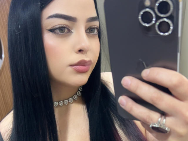 LilithAmoth, avec de longs cheveux noirs, portant un collier de diamants, prend un selfie avec un téléphone noir, a un trait d'eye-liner ailé, du rouge à lèvres rose et un anneau de nez, et regarde l'appareil photo..
