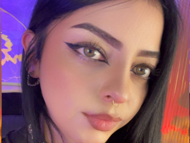 Le visage de LilithAmoth, avec ses yeux verts, son trait d'eye-liner noir en forme d'aile, ses sourcils foncés, un anneau au nez, des boucles d'oreilles dorées, ses cheveux noirs, sa peau rosée et ses lèvres roses. Son expression est légèrement moqueuse..