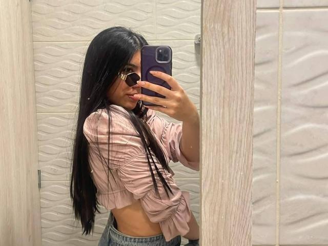 MilusSky, con lunghi capelli neri, indossa una camicia rosa annodata e occhiali, e sta scattando un selfie allo specchio con un telefono viola, mostrando il suo fianco e un accenno del suo sedere..