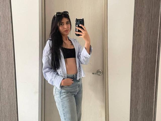 MilusSky, com cabelos longos e pretos, vestindo um sutiã preto, uma camisa azul clara e jeans de cintura alta, tirando uma selfie no espelho, mostrando um rosto fofo e com a mão no bolso..