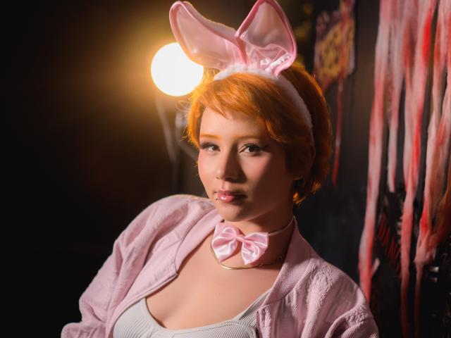 ChloeeWilliams with short red hair, pink bunny ears, pink bowtie, and pink jacket, has puffy tits and a cute face, illuminated by a bright light source.