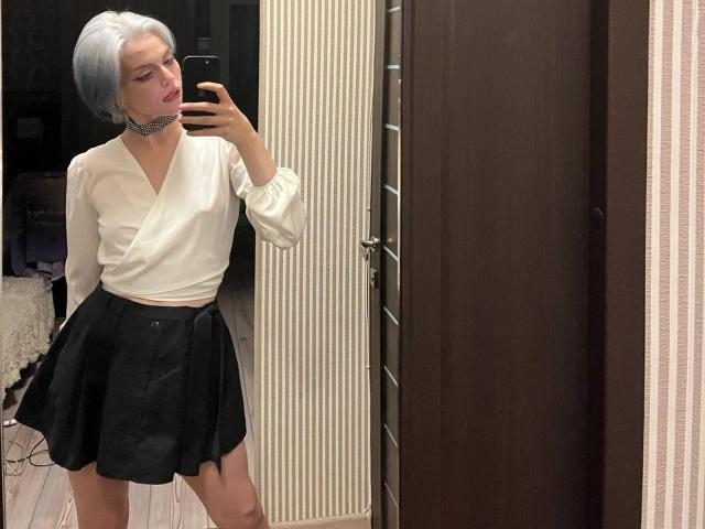 HanhFontillas, con i capelli corti e grigi, una camicetta bianca, una gonna nera e una collana, sta scattando un selfie allo specchio, mostrando il sedere e un piccolo gluteo carino in una stanza elegante e moderna..