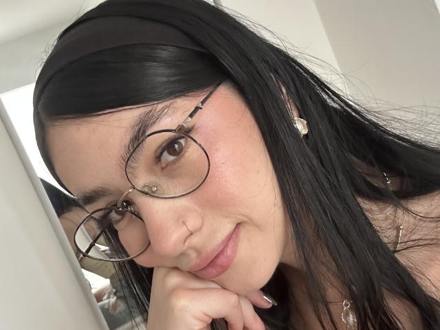 EvaaFarix with black hair, glasses, nose ring, and heart earrings, smiling slightly, resting chin on hand, wearing a black headband. She has a cute, pretty face.